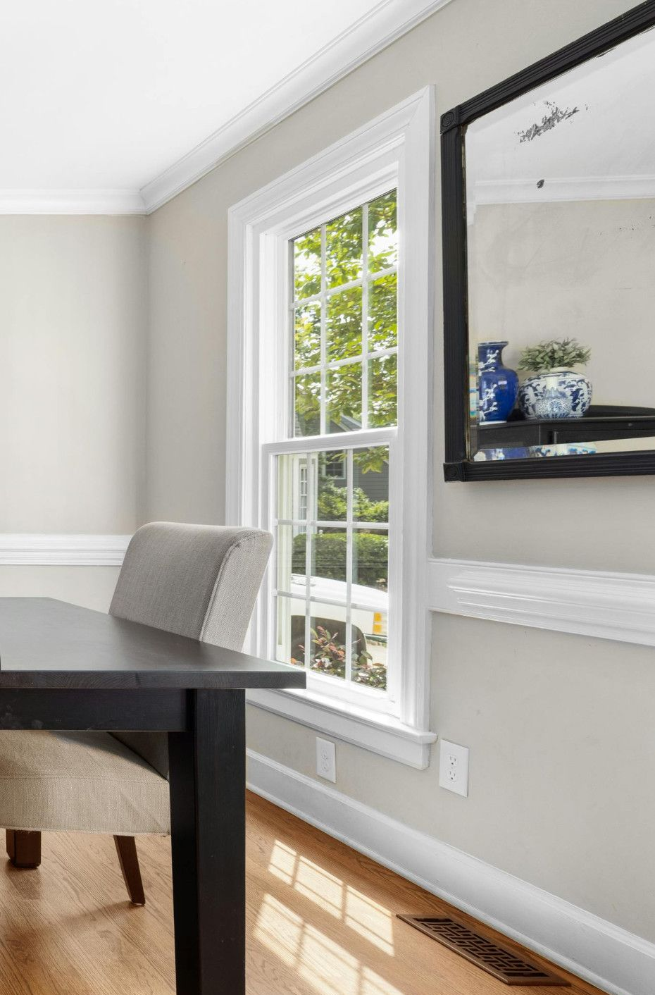 A dining room scene with a window, table, chair, and mirror. The room has light gray walls and wood floors.