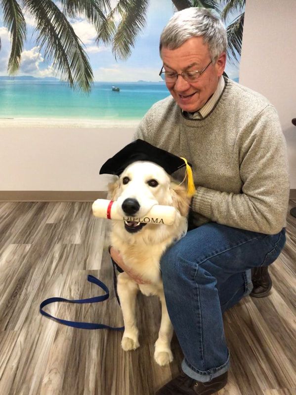 A man is kneeling down next to a dog wearing a graduation cap and holding a diploma.