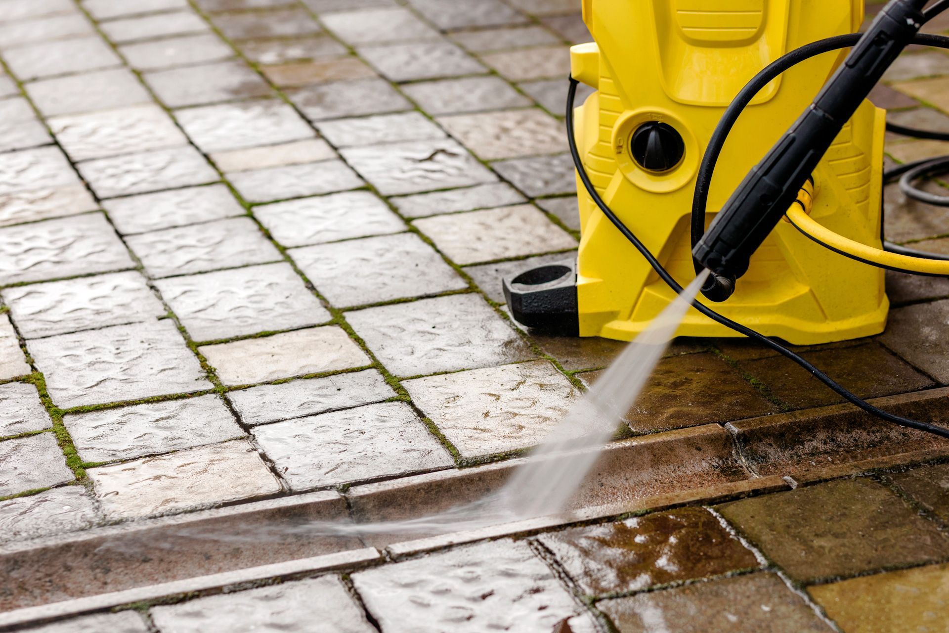 A yellow high pressure washer is cleaning a brick sidewalk.