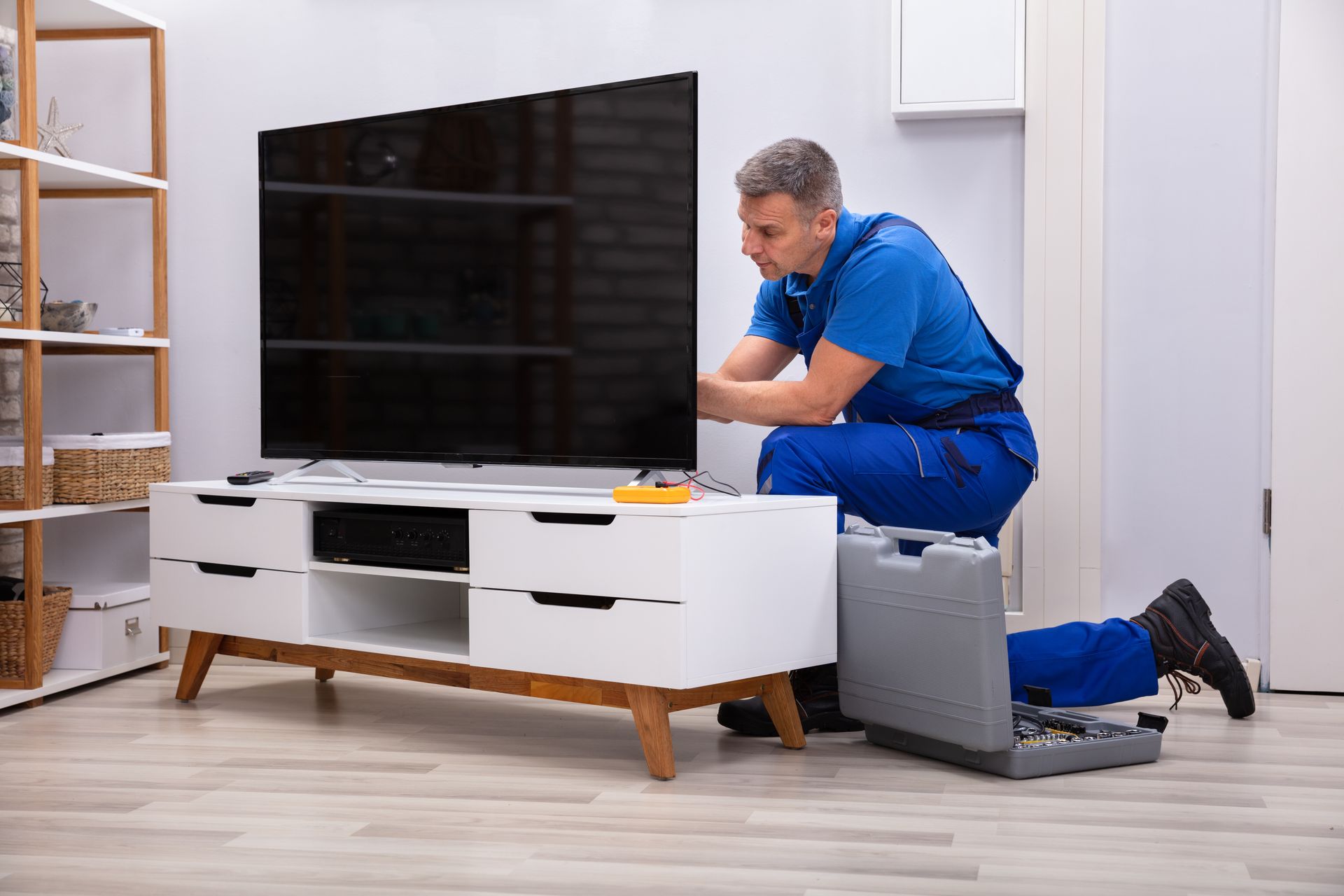 Man in blue jumpsuit kneeling, fixing a TV on a white cabinet, tools nearby. Interior setting.