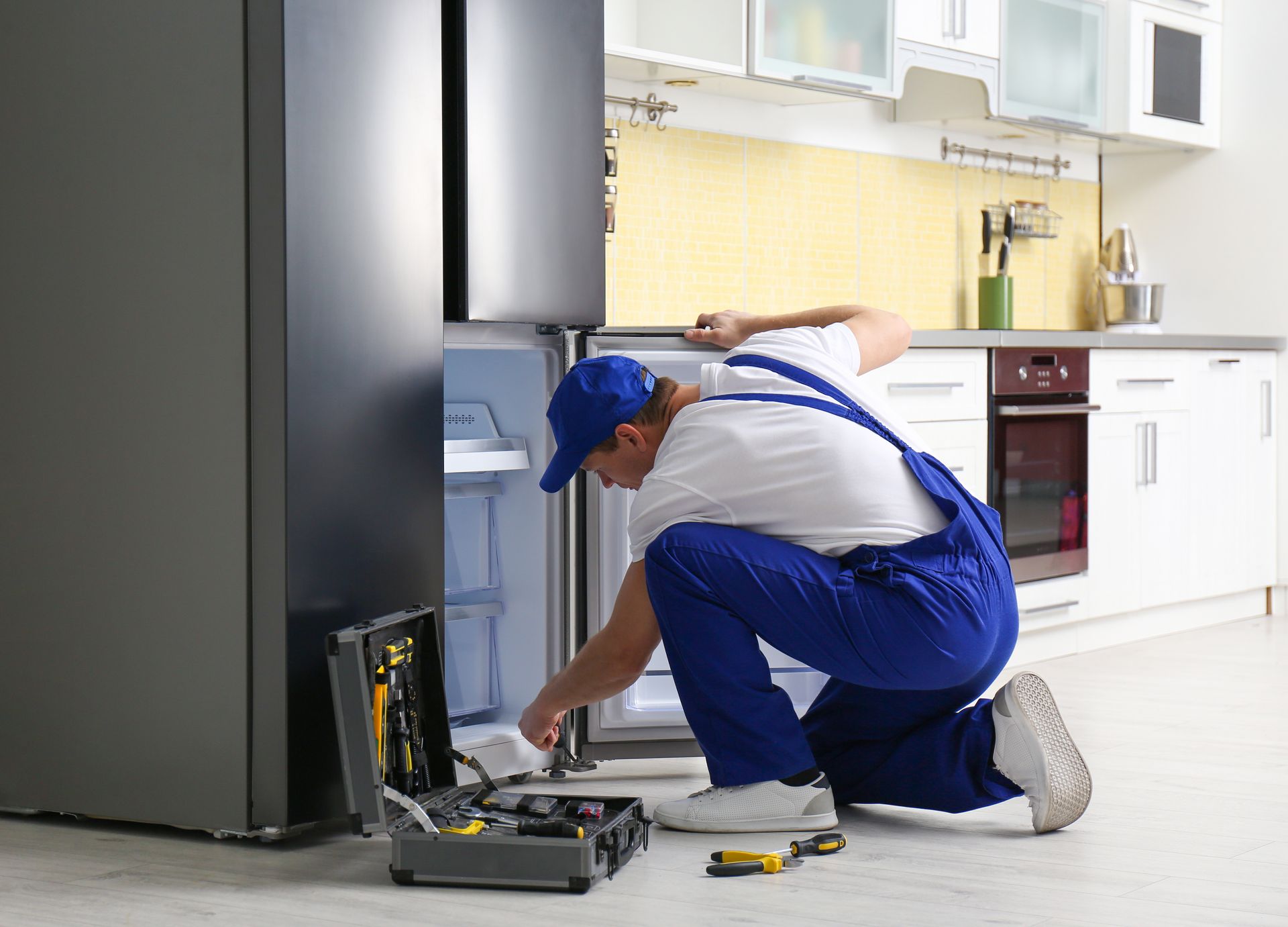 A repairman in blue overalls works on a refrigerator in a bright white kitchen.