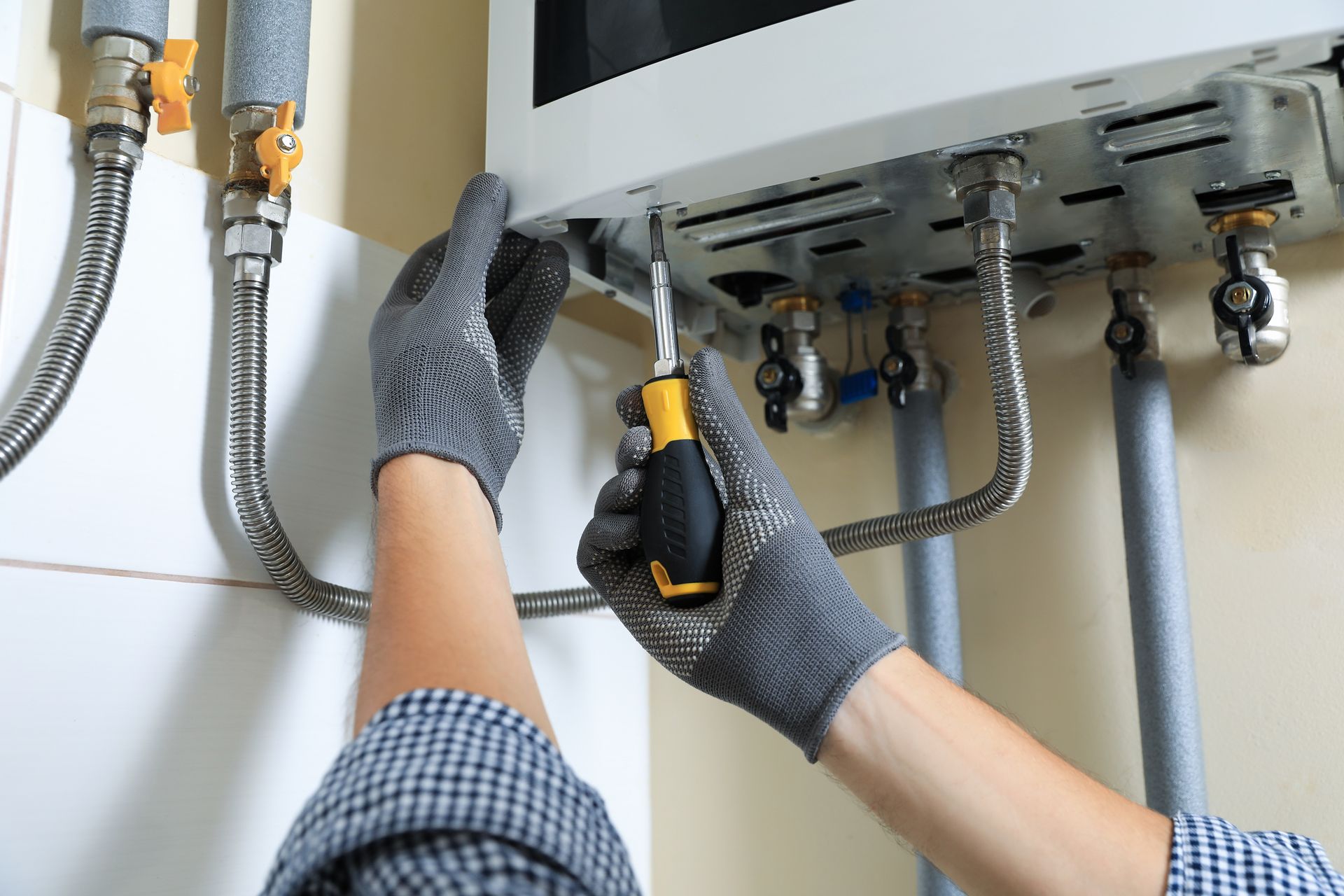 Person wearing gloves uses a screwdriver to work on a white wall-mounted appliance with pipes.