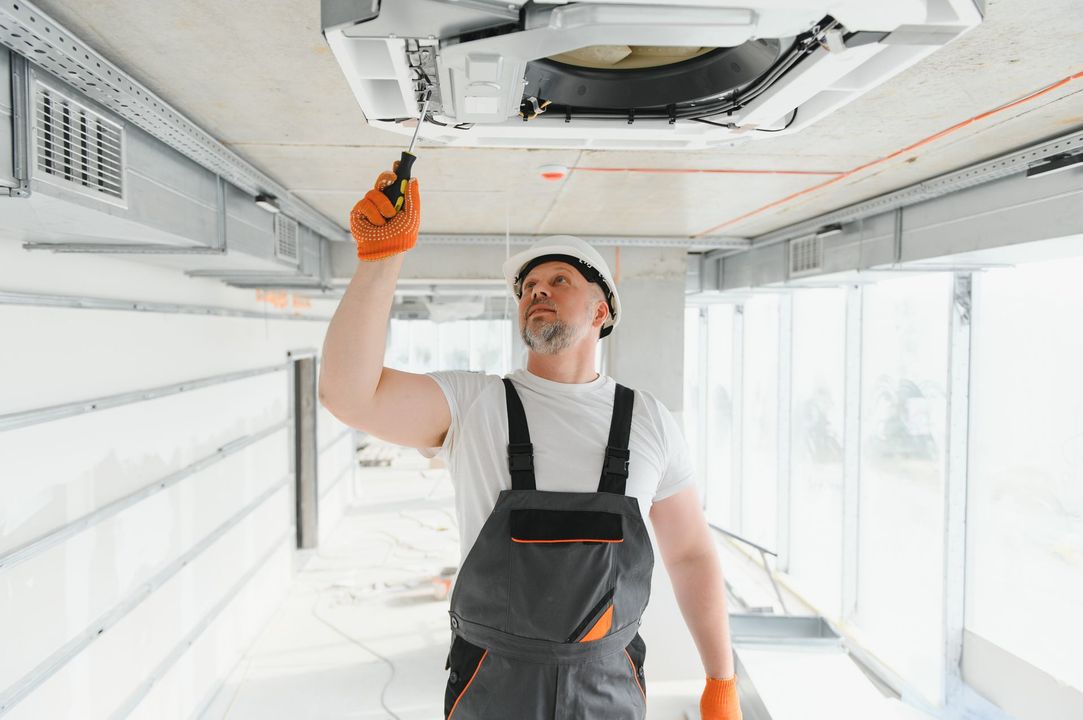 Man in overalls, helmet, and gloves working on ceiling air conditioning unit in a construction setting.