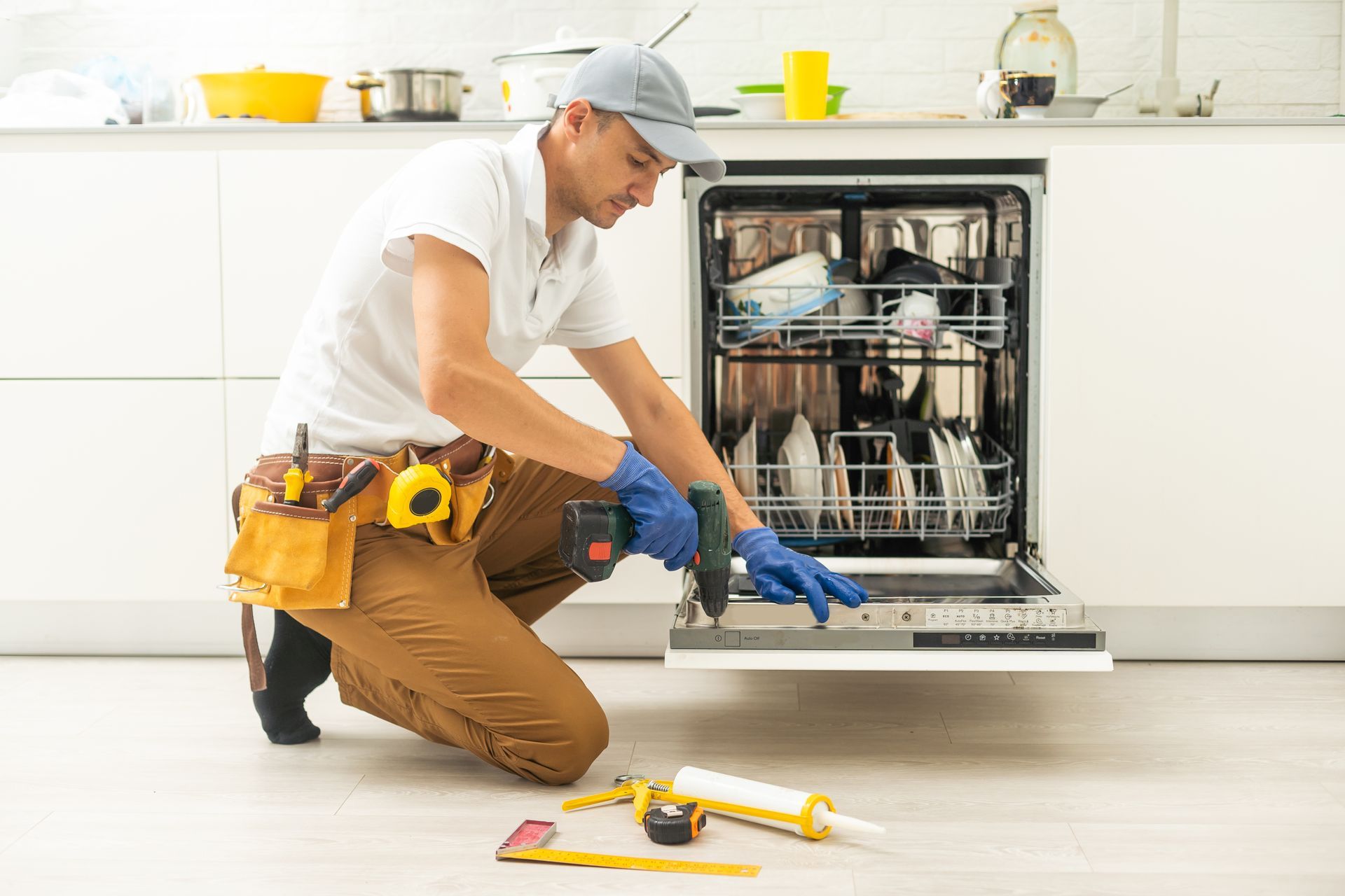 Man in work clothes fixing a dishwasher in a kitchen, holding a drill and surrounded by tools.