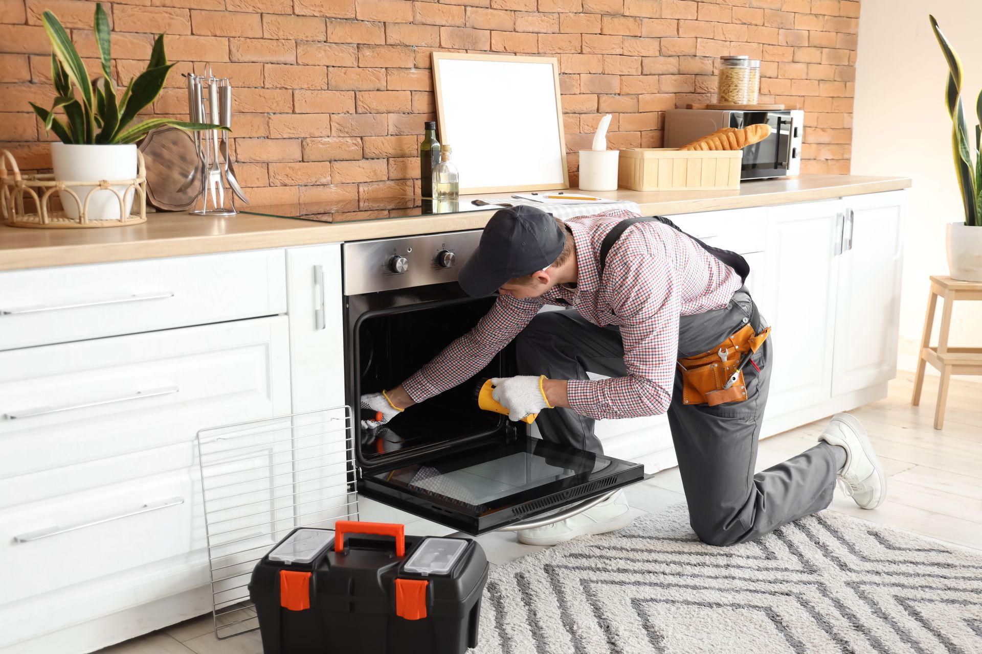 A repairman kneels, inspecting an oven in a kitchen. He wears a cap, tools visible, and the oven door is open.