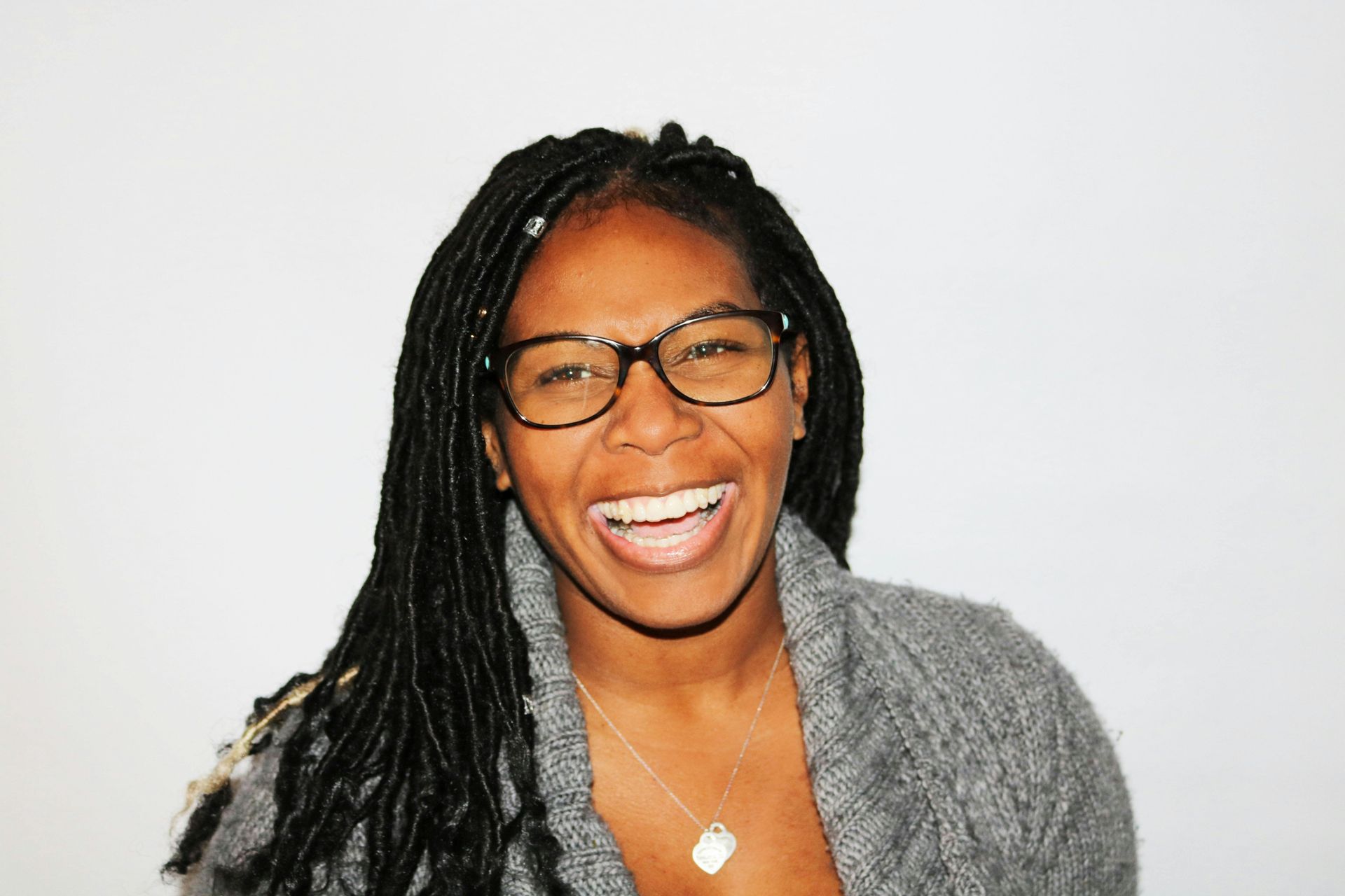 Woman wearing glasses laughs, hair in dreadlocks, wearing a gray sweater, against a white background.