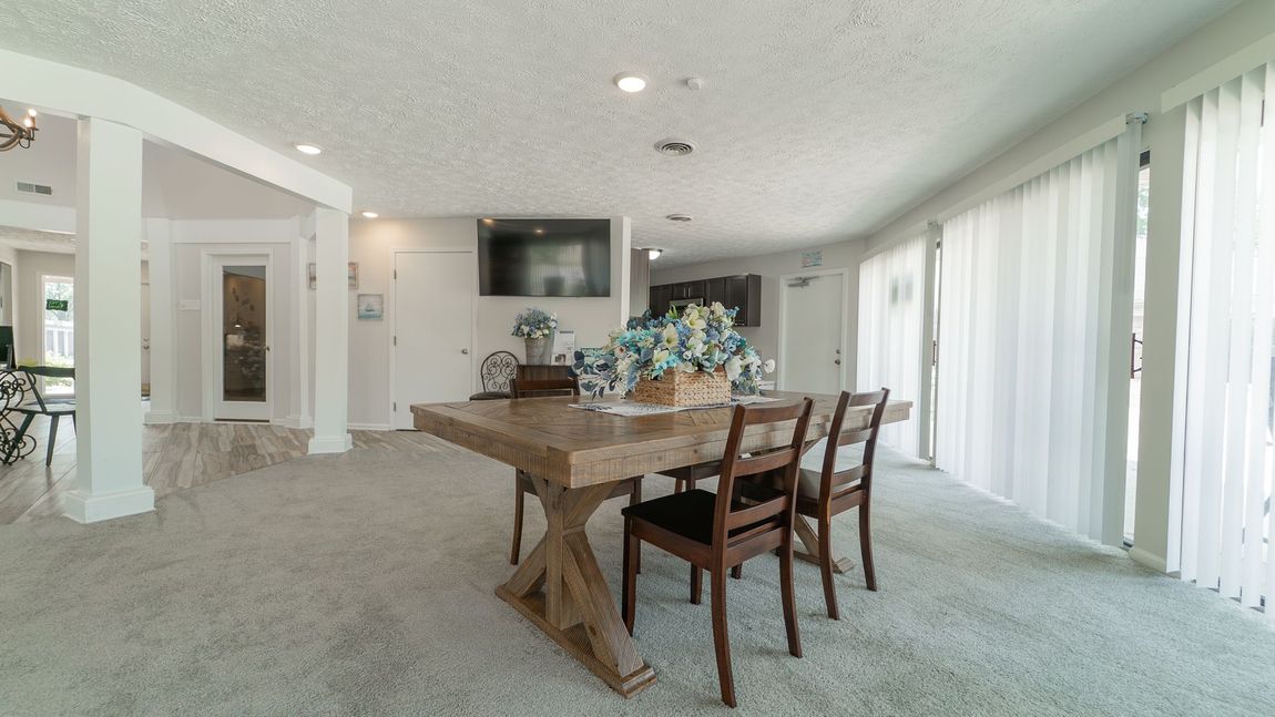 Spacious dining area with beige carpet, large wooden table, chairs, and window curtains. TV mounted on the wall.