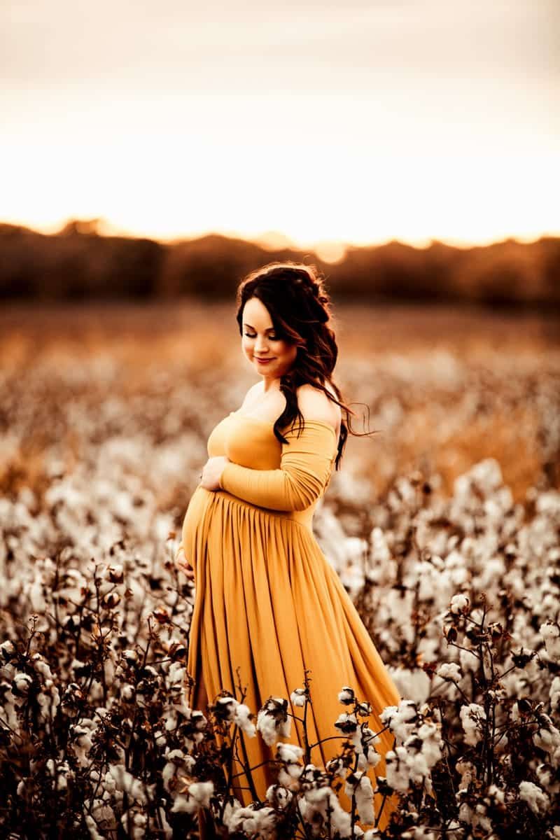 A pregnant woman in a yellow dress is standing in a field of cotton flowers.