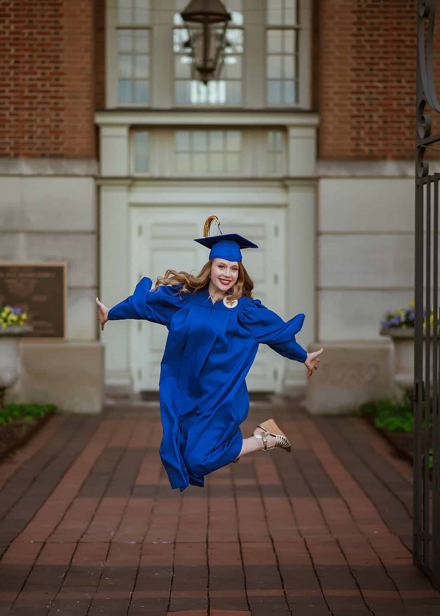 A woman in a blue graduation cap and gown is jumping in the air.