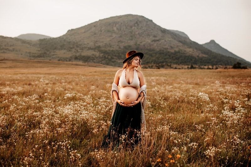 A pregnant woman is standing in a field of flowers with a mountain in the background.