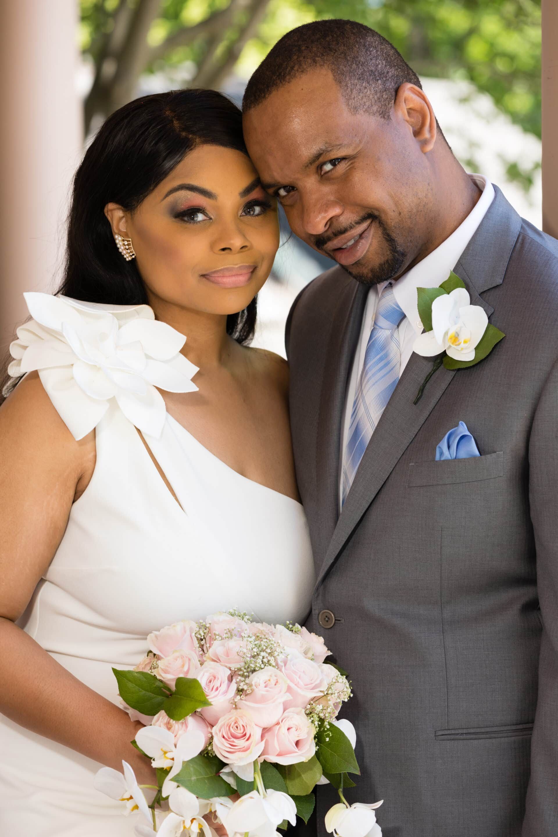 The bride and groom are posing for a picture and the bride is holding a bouquet of flowers.