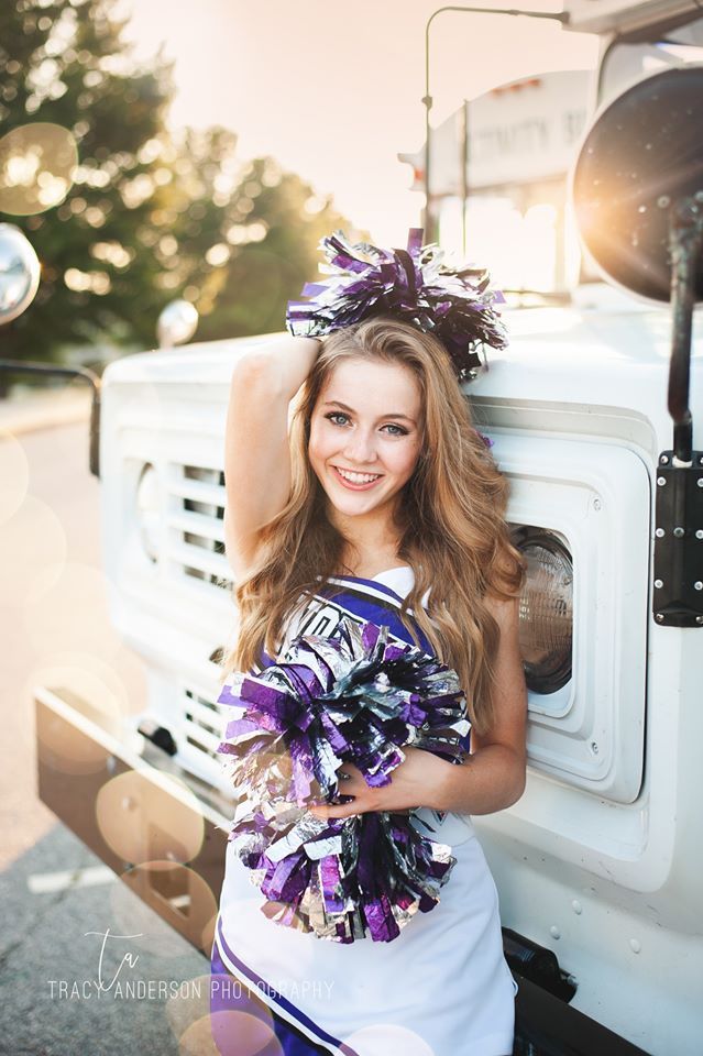 A cheerleader is standing in front of a white truck holding pom poms.