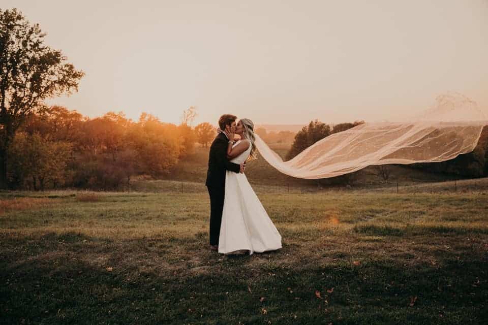 A bride and groom kissing in a field with a veil blowing in the wind.