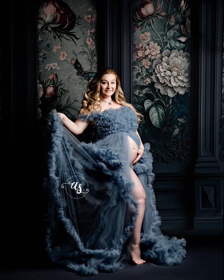 A pregnant woman in a blue dress is standing in front of a floral wall.