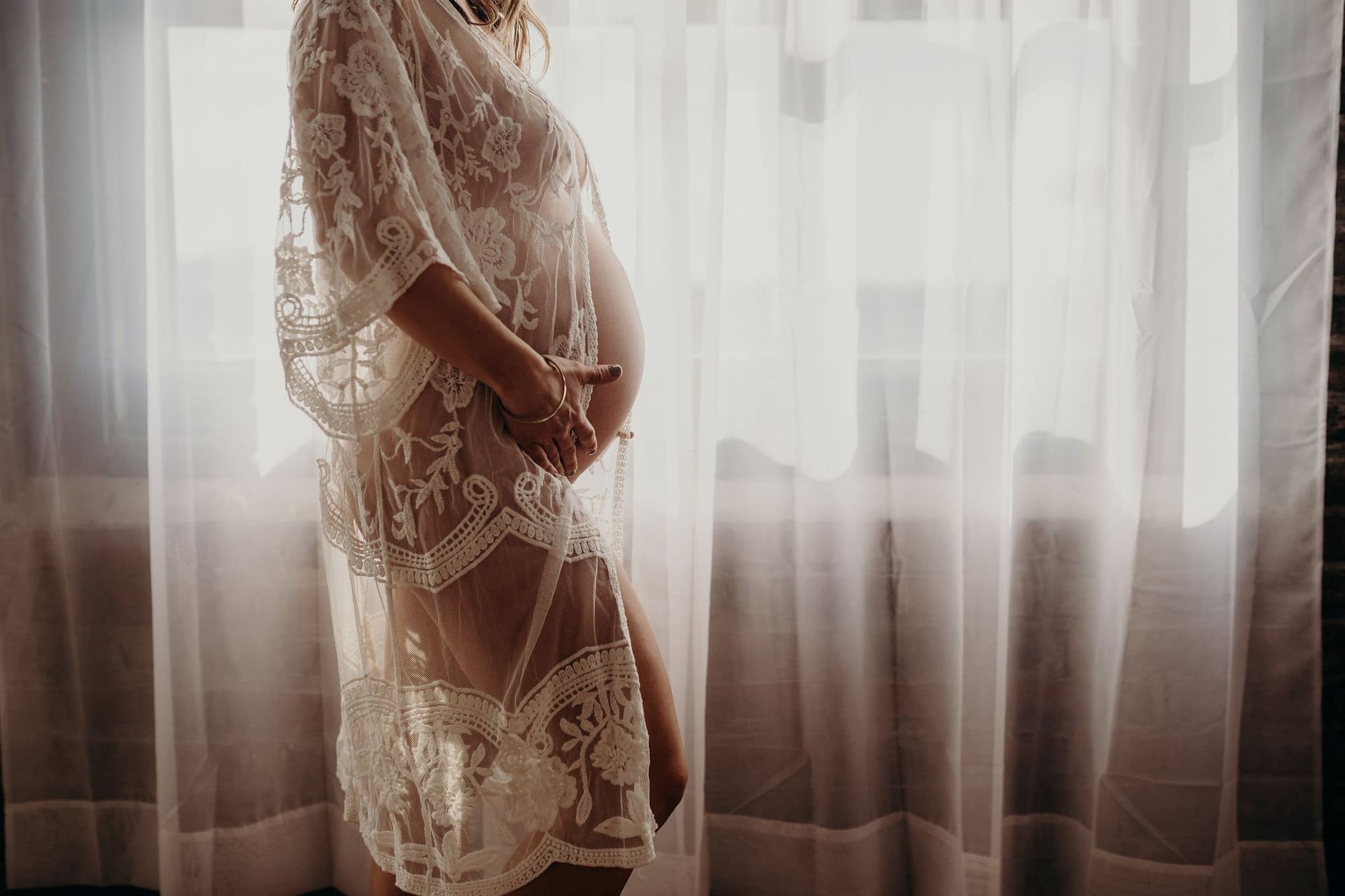 A pregnant woman in a white lace dress is standing in front of a window.