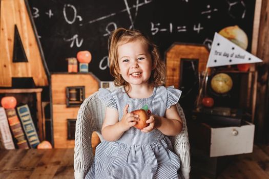 A little girl is sitting in a chair holding an apple in front of a blackboard.