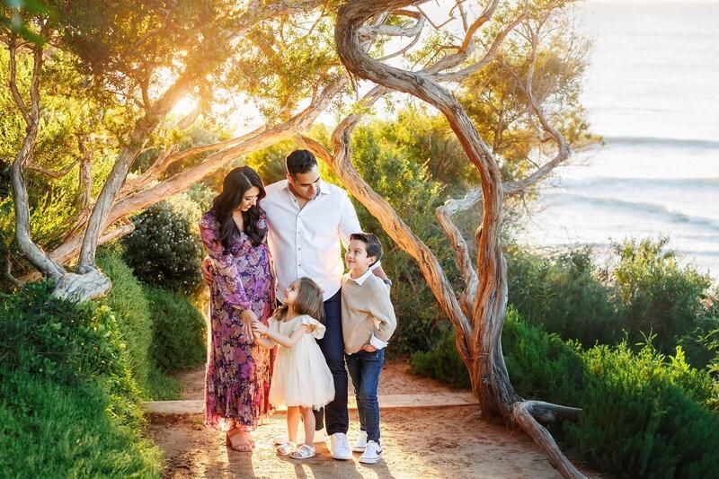 A family is walking under a tree on a path near the ocean.