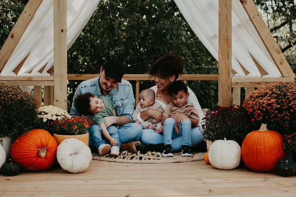 A family is sitting on a porch surrounded by pumpkins and flowers.