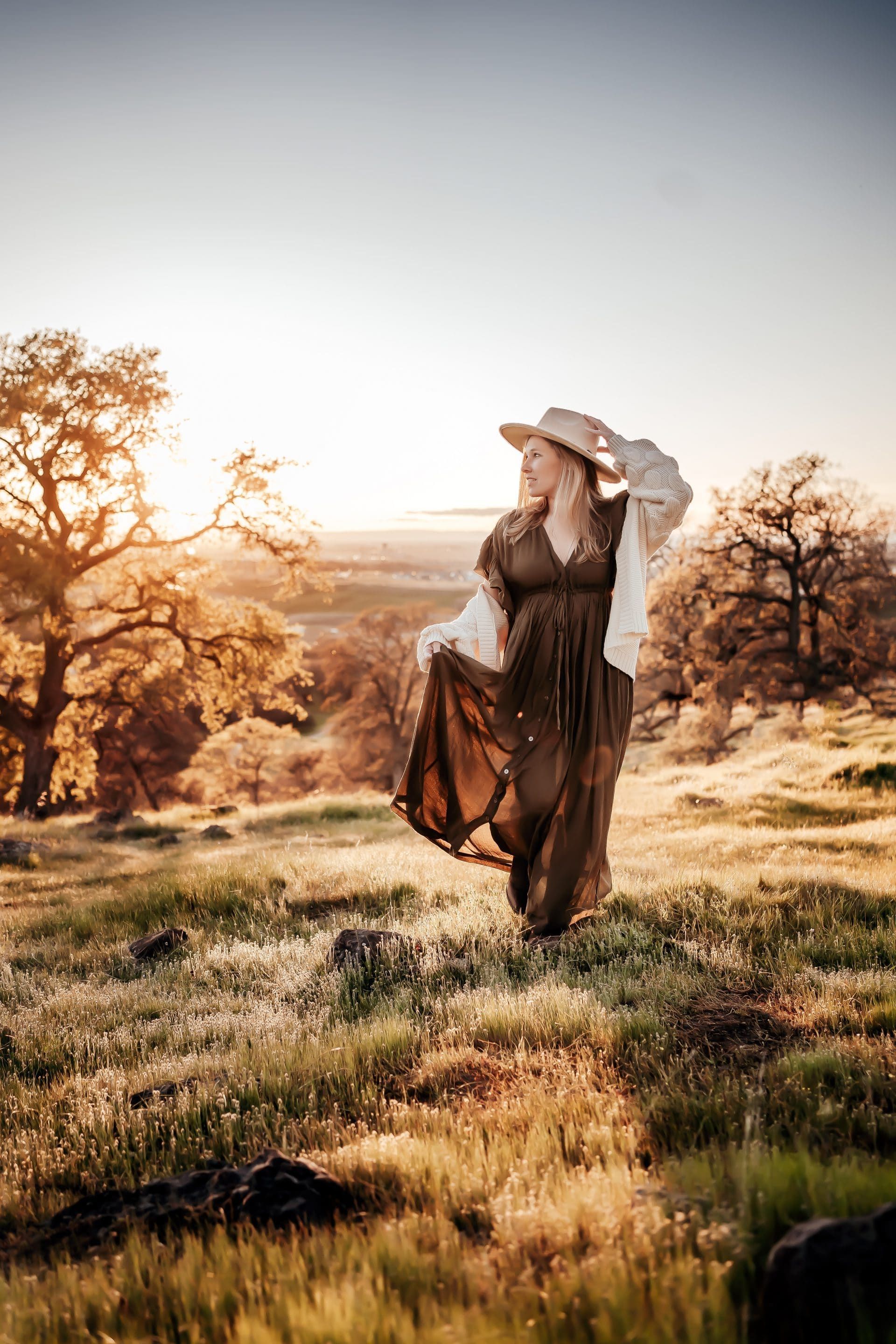 A woman in a long dress and hat is standing in a field at sunset.