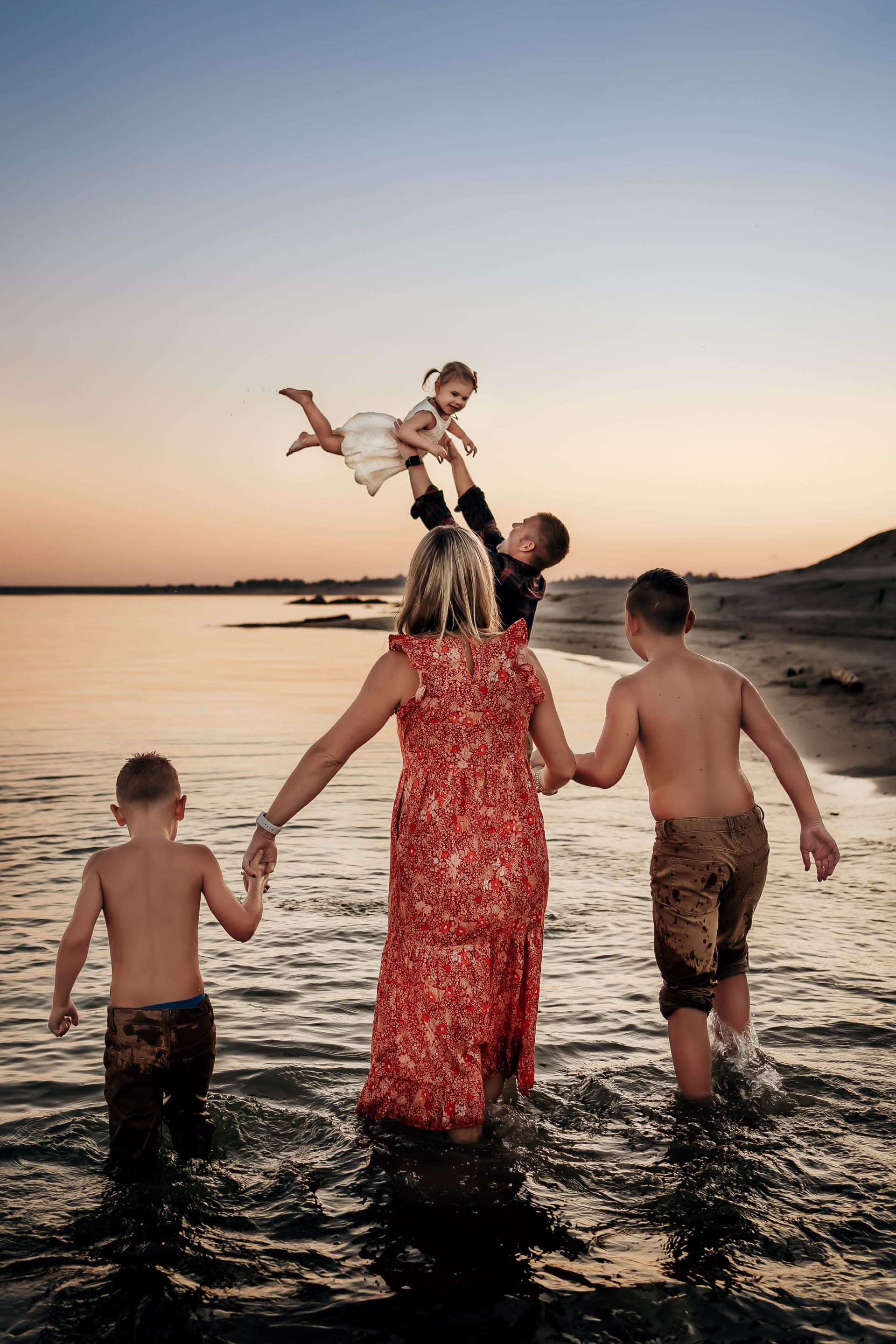 A woman is holding a child in the air while two boys are playing in the water.