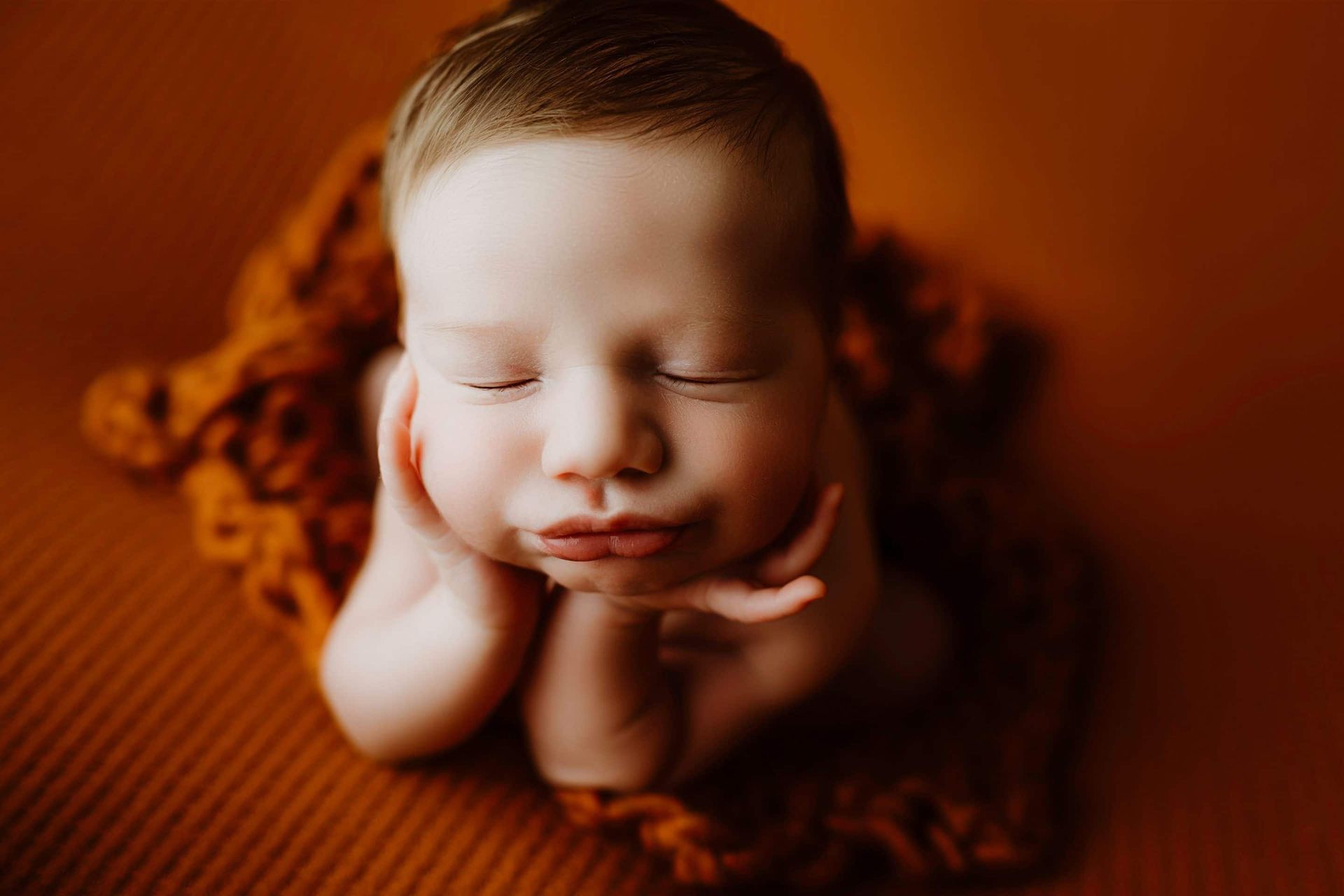 A newborn baby is sleeping on a blanket with his hands on his face.