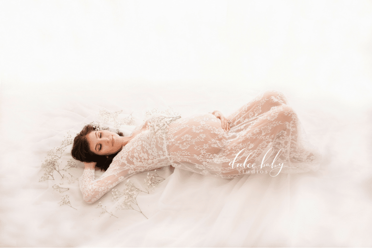 A pregnant woman in a white lace dress is laying on a bed.