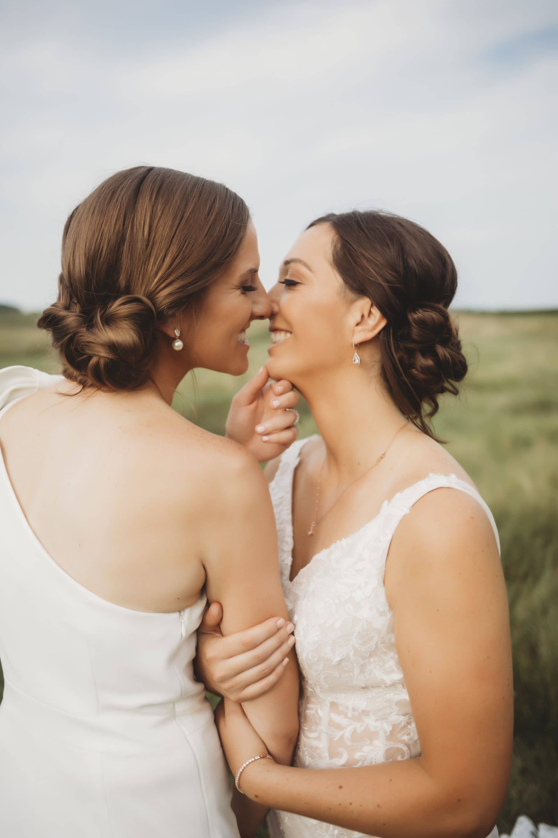 Two women in white dresses are kissing in a field.