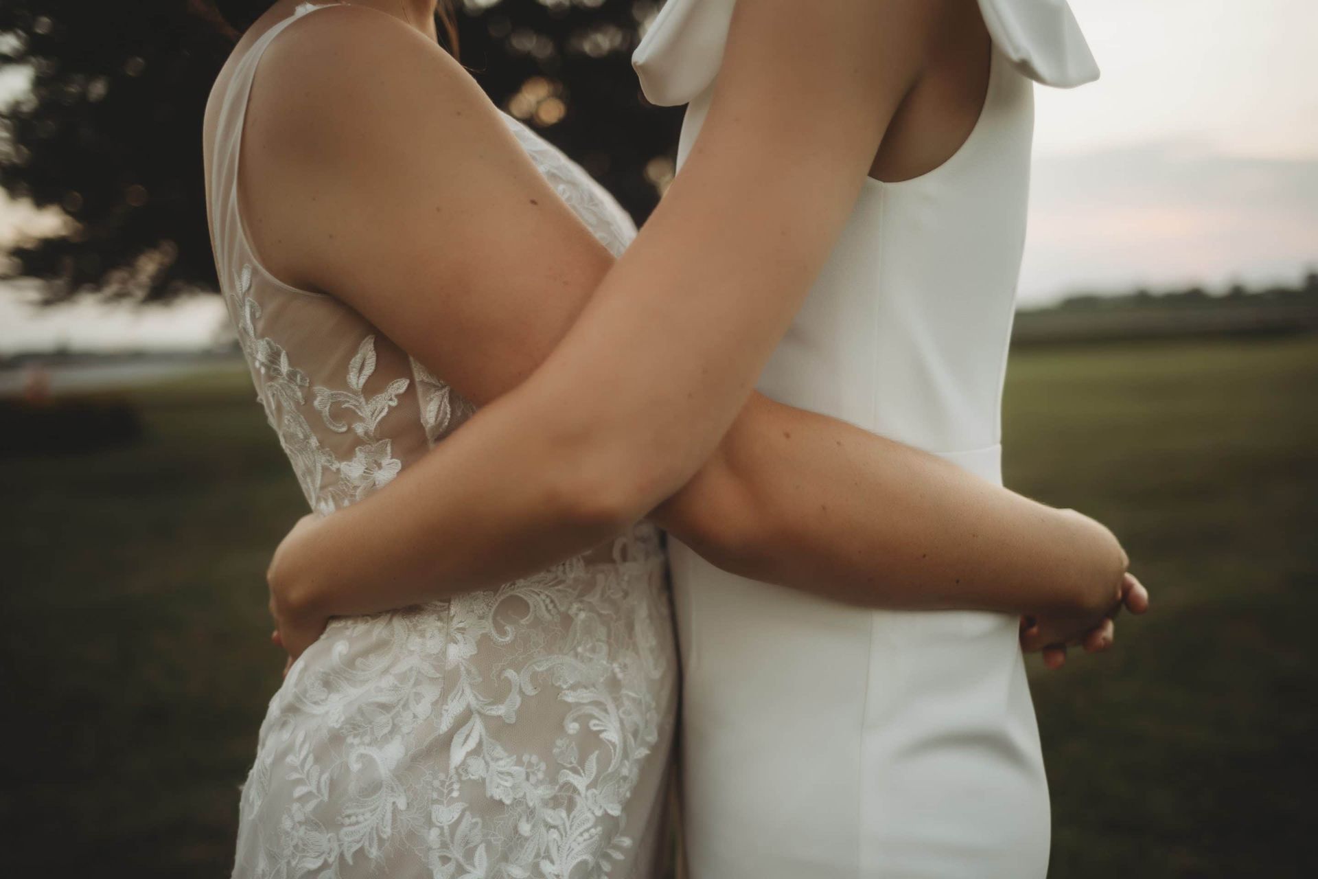 Two women in white dresses are hugging each other in a field.