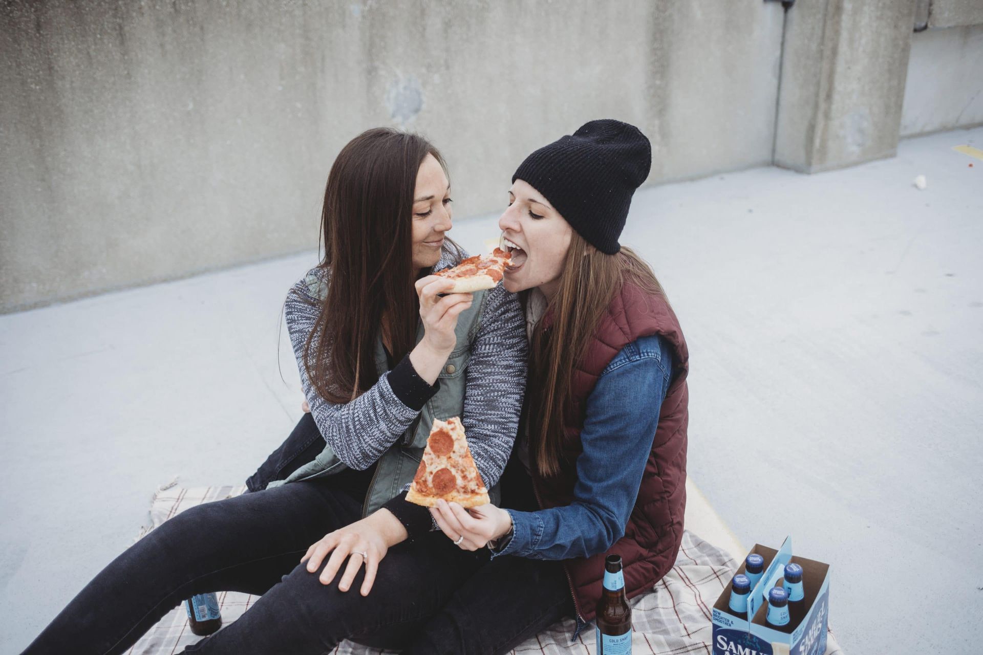 Two women are sitting on a blanket eating pizza and drinking beer.