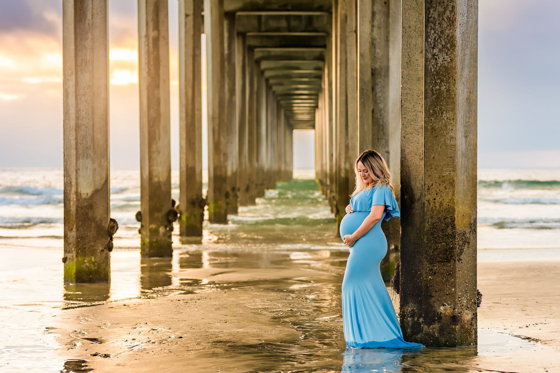 A pregnant woman in a blue dress is standing under a pier on the beach.