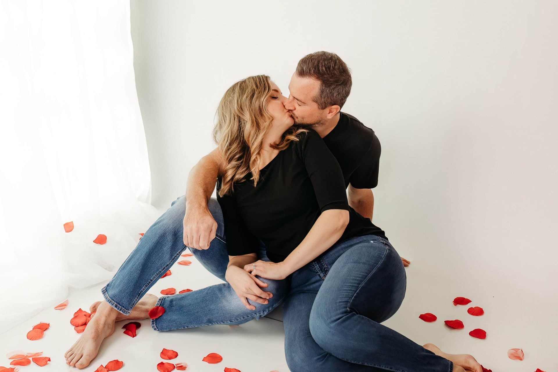 A man and a woman are kissing while sitting on the floor surrounded by rose petals.