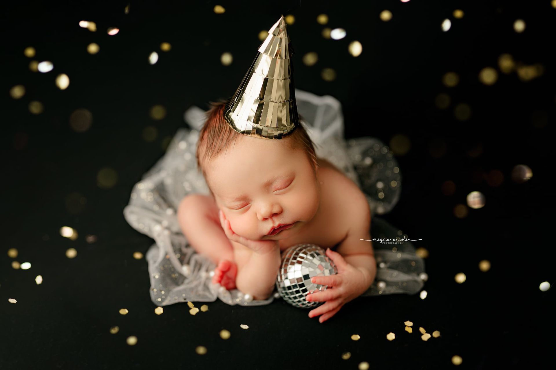 A newborn baby wearing a party hat is holding a disco ball.