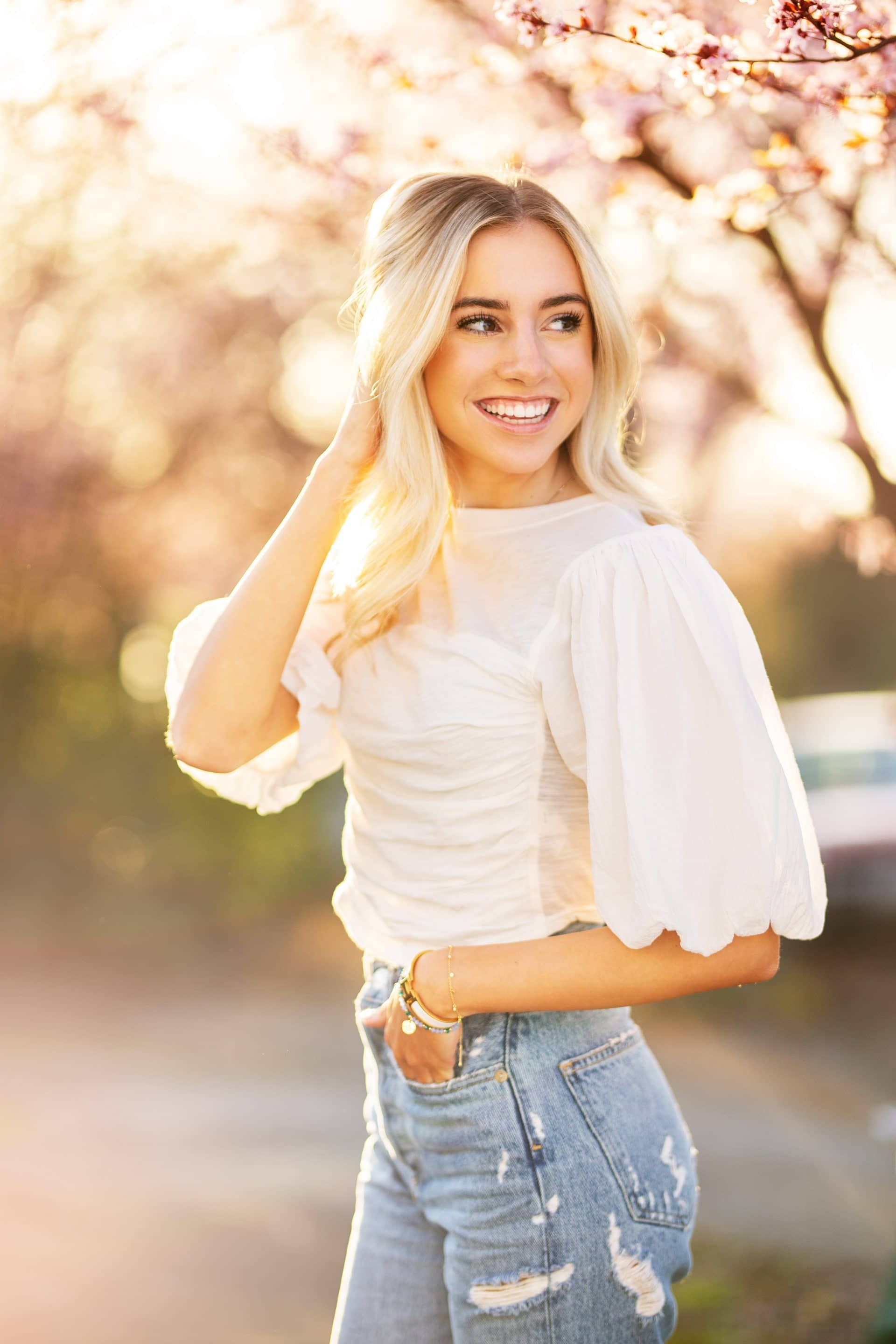 A woman in a white top and jeans is standing under a cherry blossom tree.