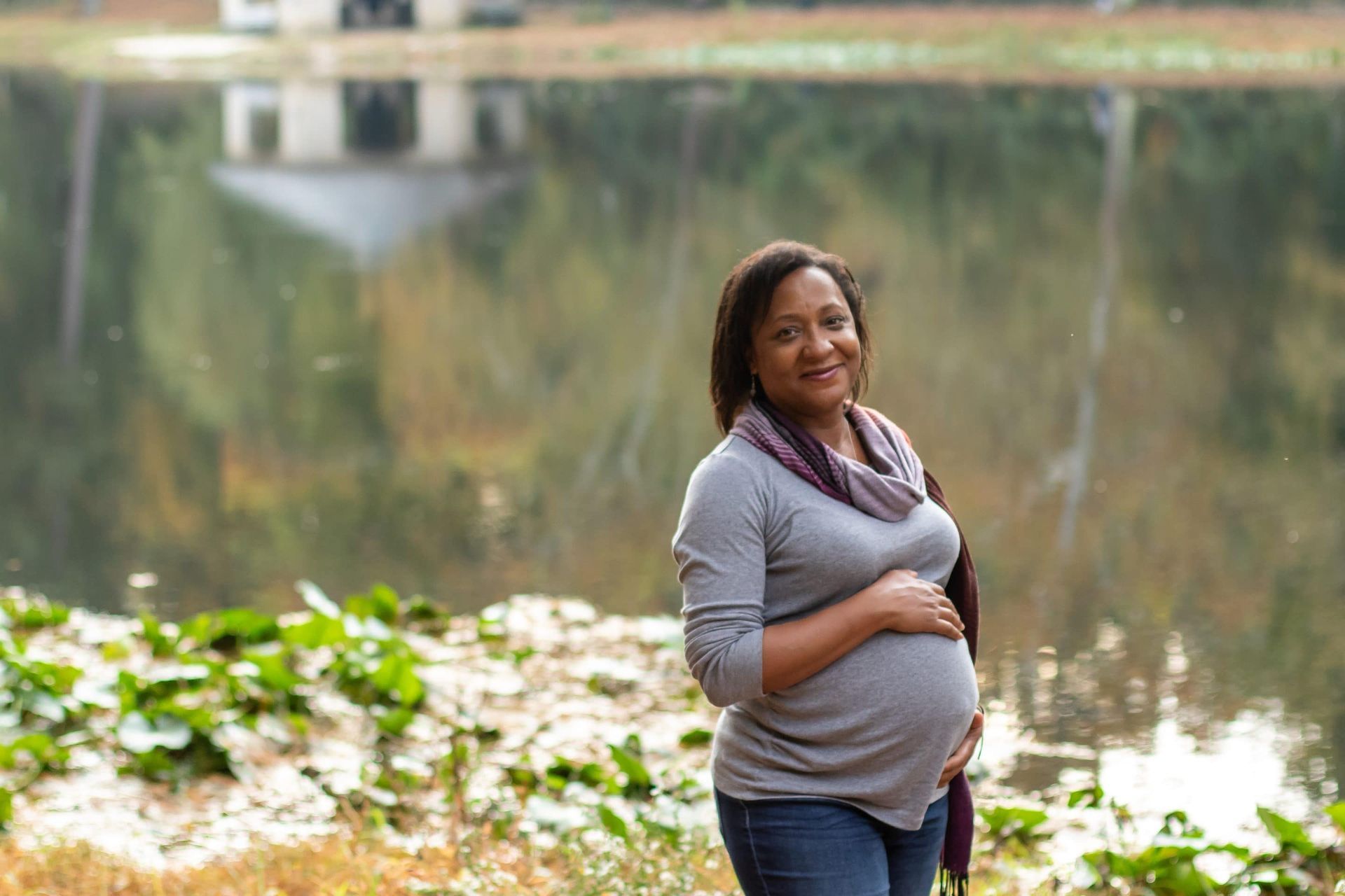 A pregnant woman is standing in front of a lake holding her belly.