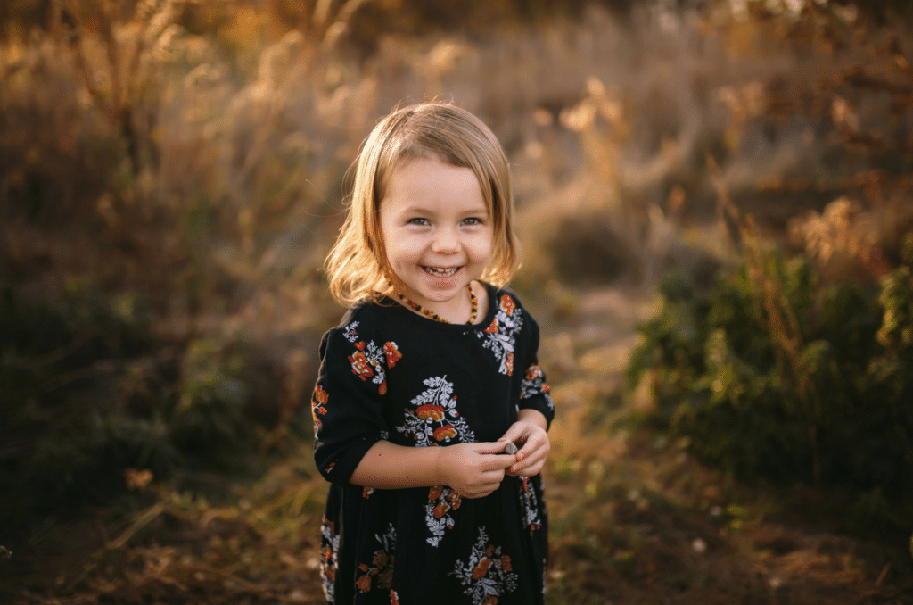 A little girl in a black dress is standing in a field and smiling.