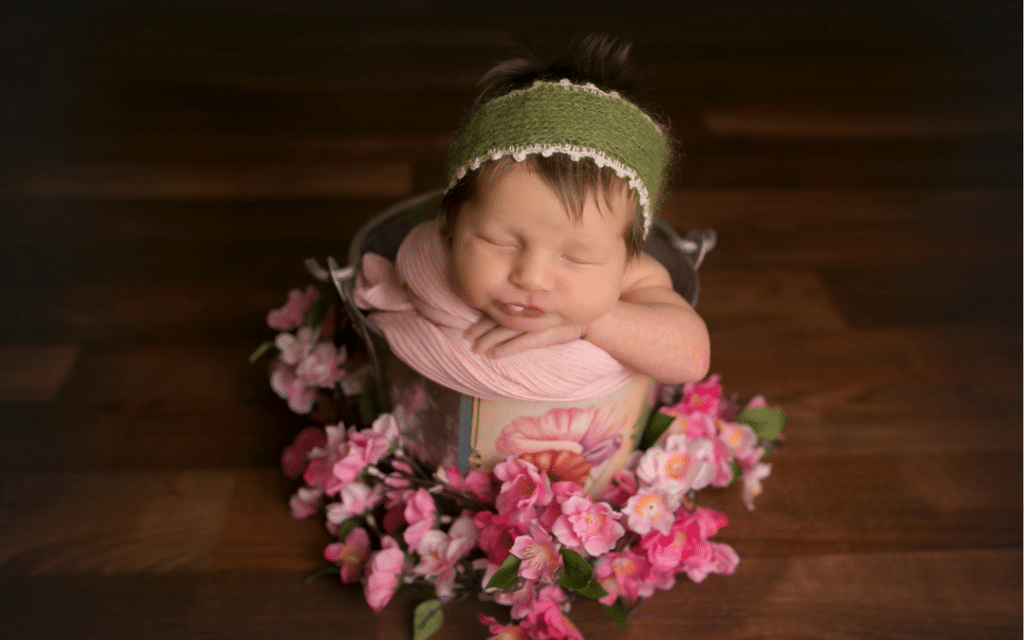 A newborn baby is sleeping in a bucket surrounded by pink flowers.