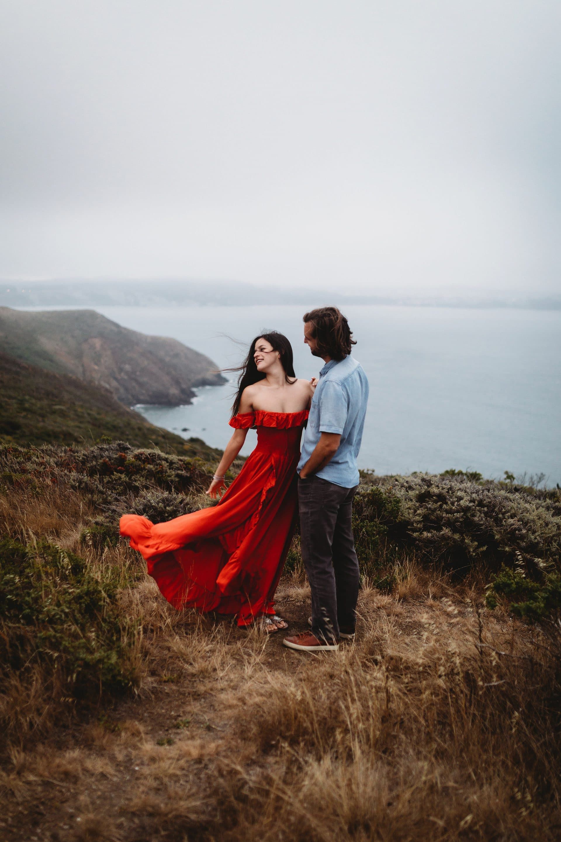 A man and a woman are standing on top of a hill overlooking the ocean . the woman is wearing a red dress.