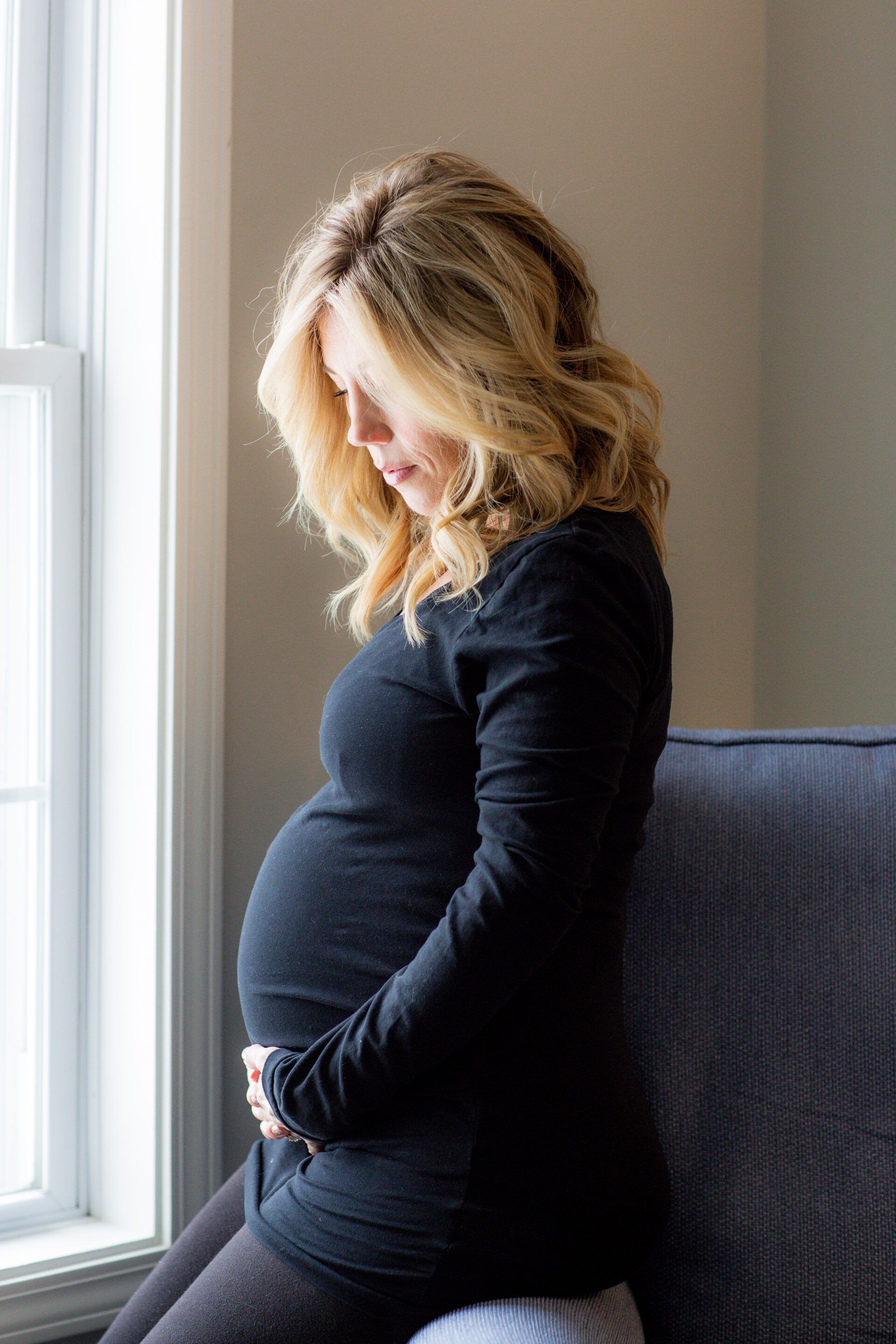 A pregnant woman is sitting on a couch in front of a window holding her belly.