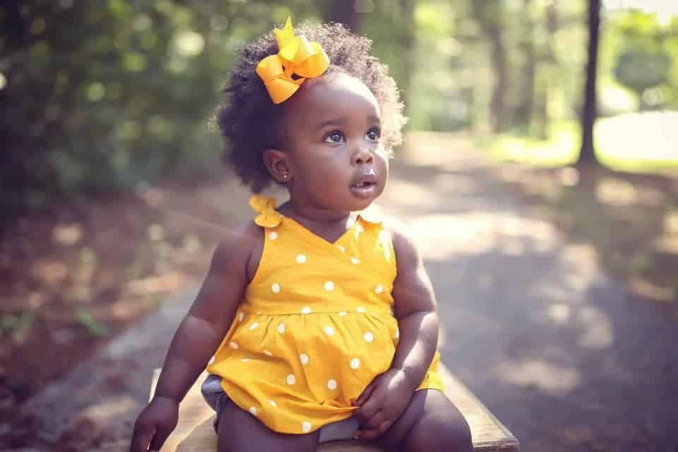 A baby girl wearing a yellow dress and a yellow bow is sitting on a bench.