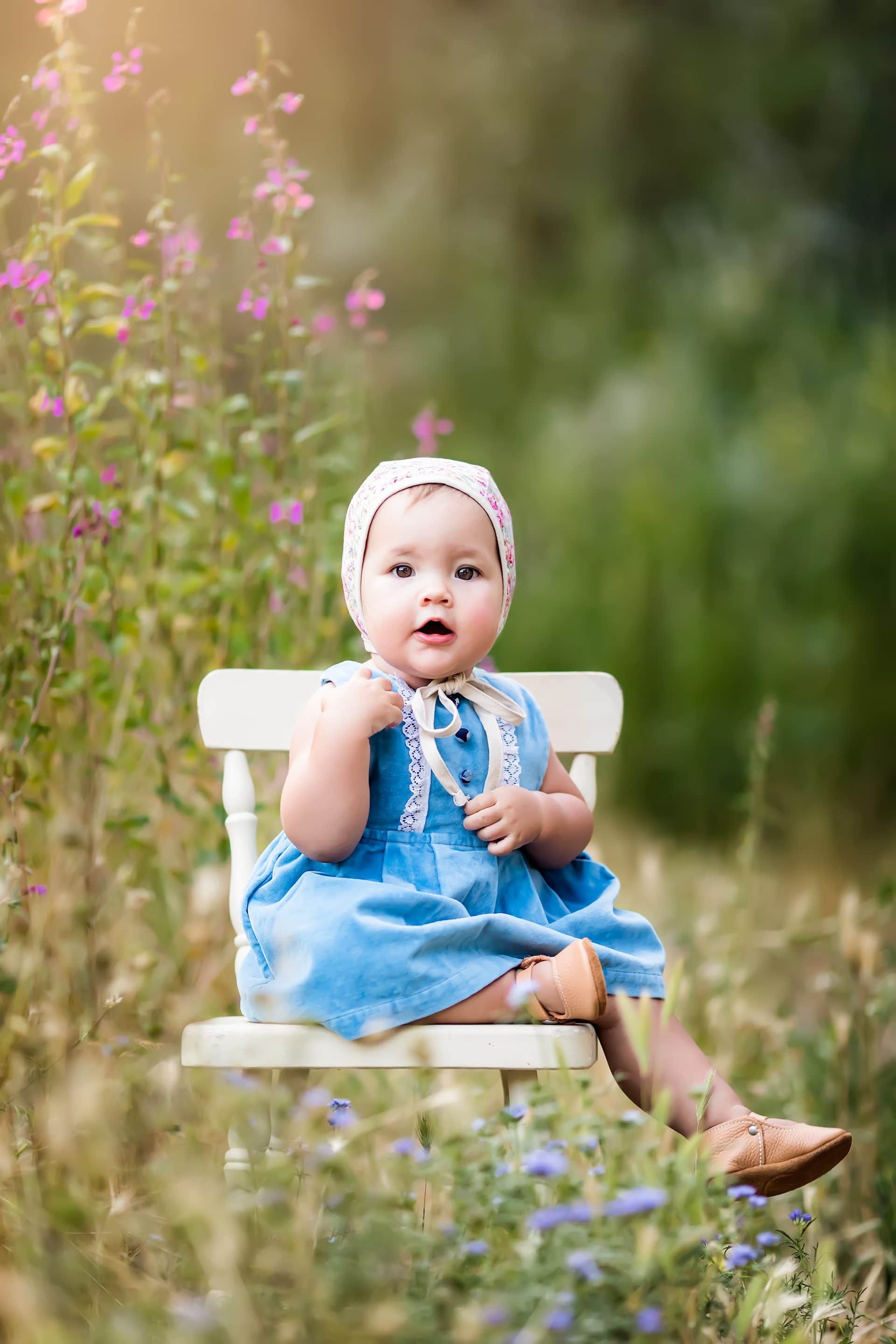 A baby in a blue dress is sitting on a white bench in a field of flowers.