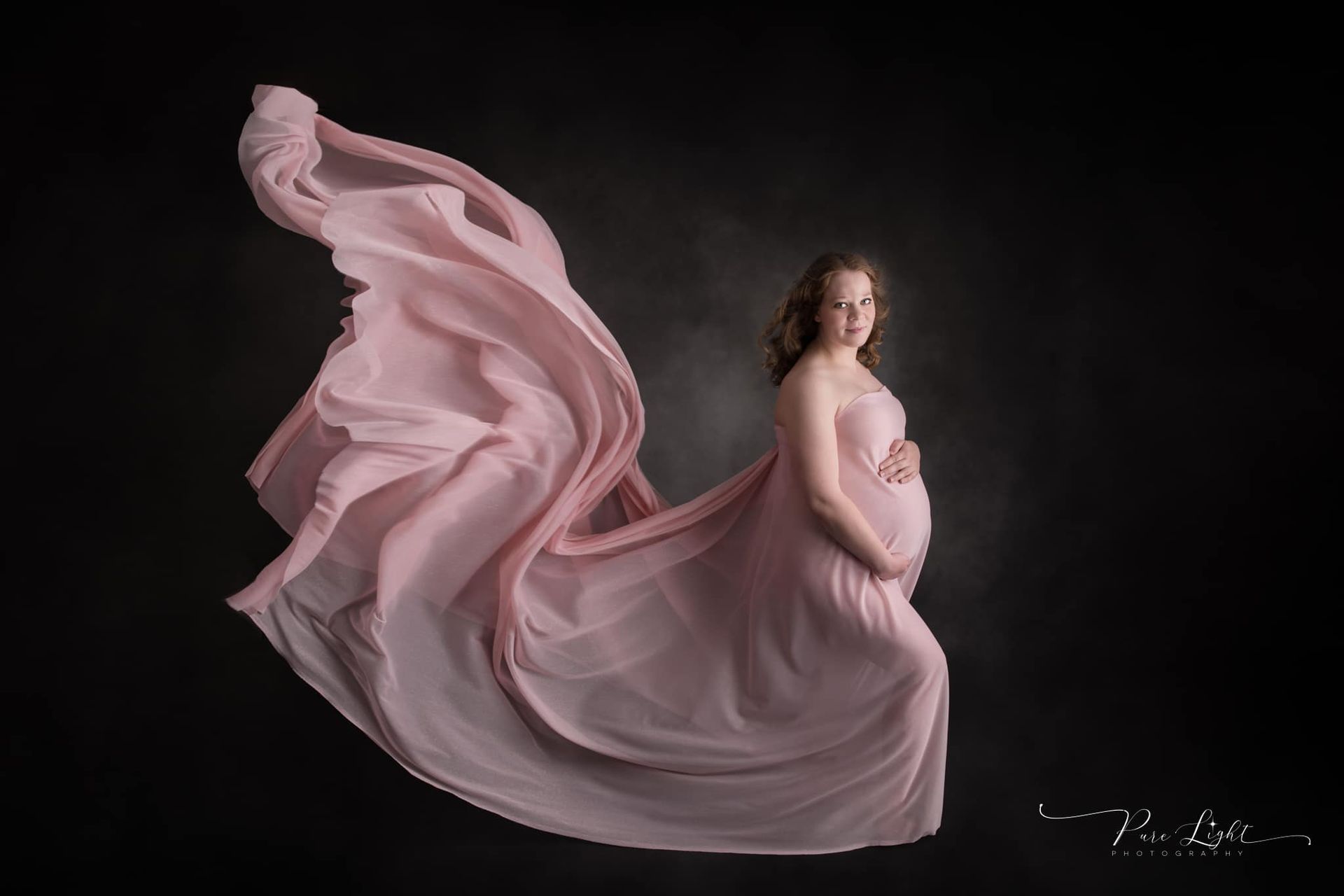 A pregnant woman in a long pink dress is standing in front of a black background.