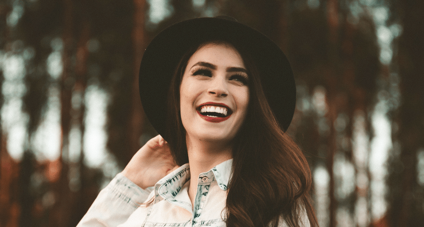 A woman wearing a hat is smiling in front of a forest.