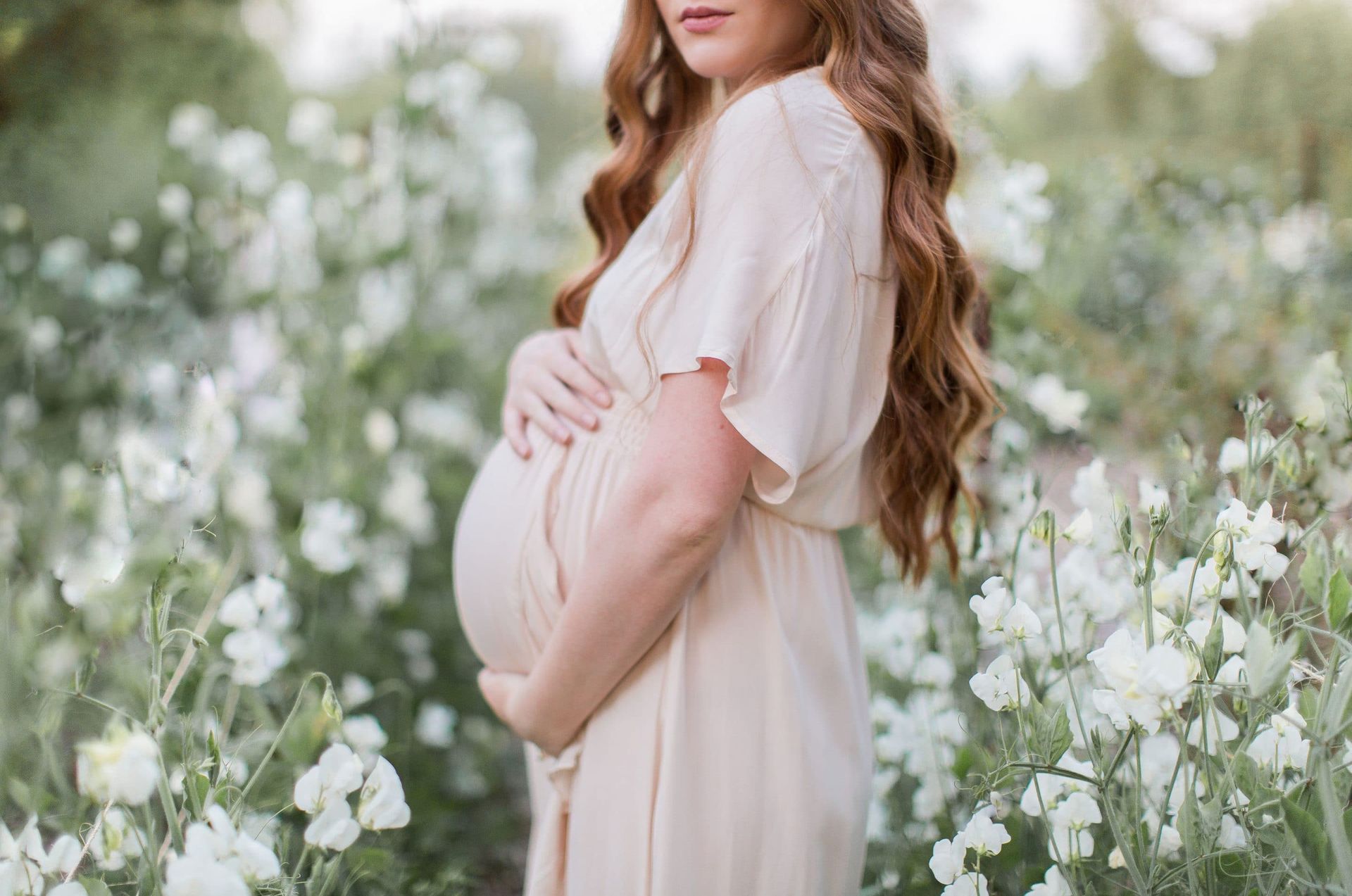 A pregnant woman in a white dress is standing in a field of white flowers.