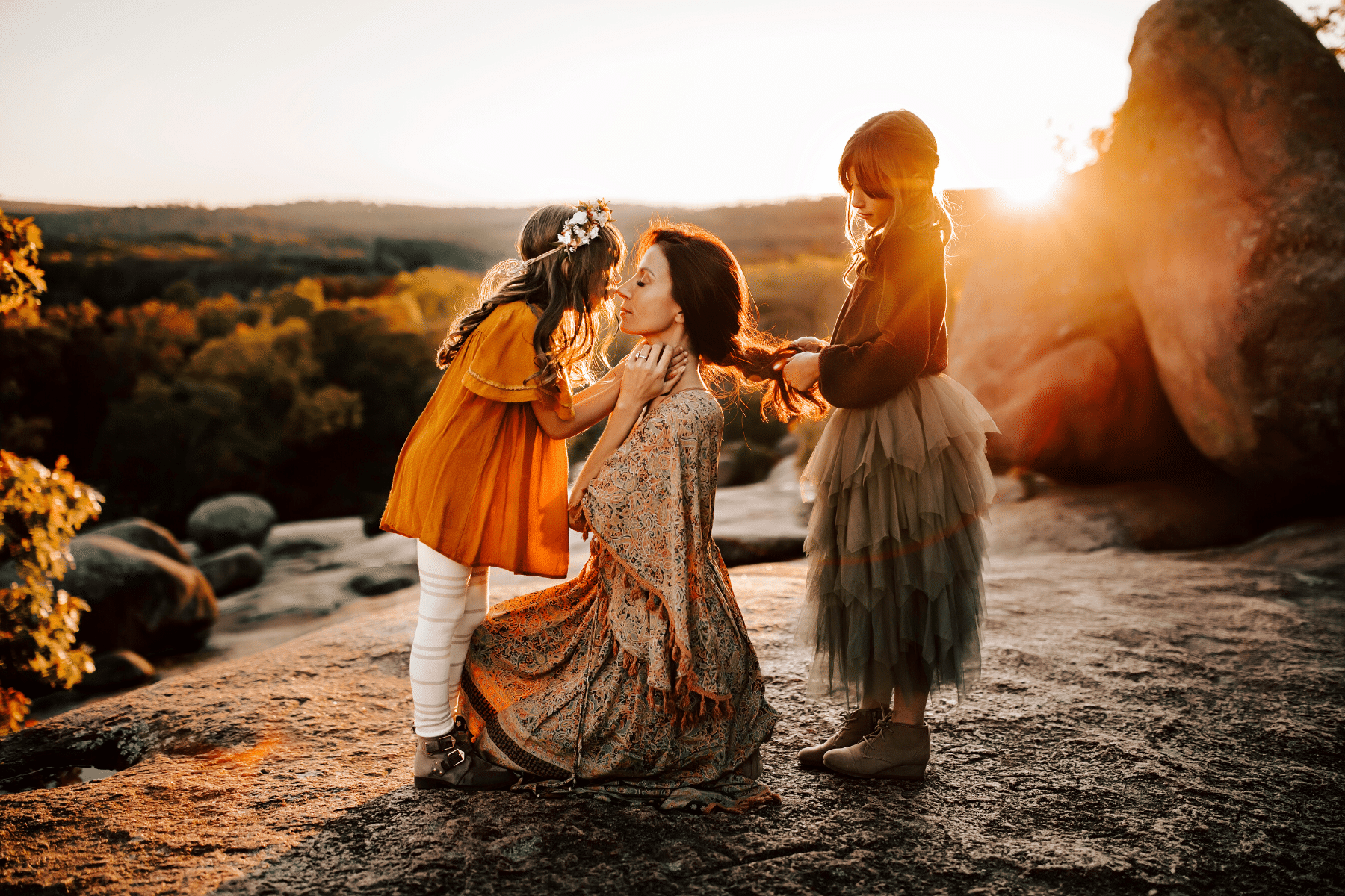 A woman is kneeling down with two little girls playing with her hair.