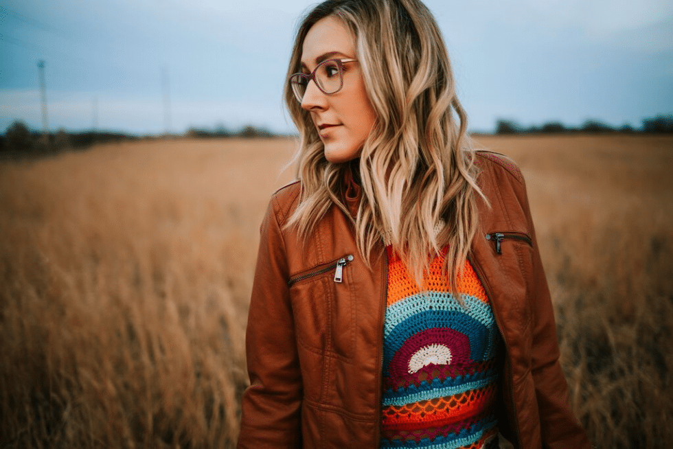 A woman wearing glasses and a leather jacket is standing in a field.