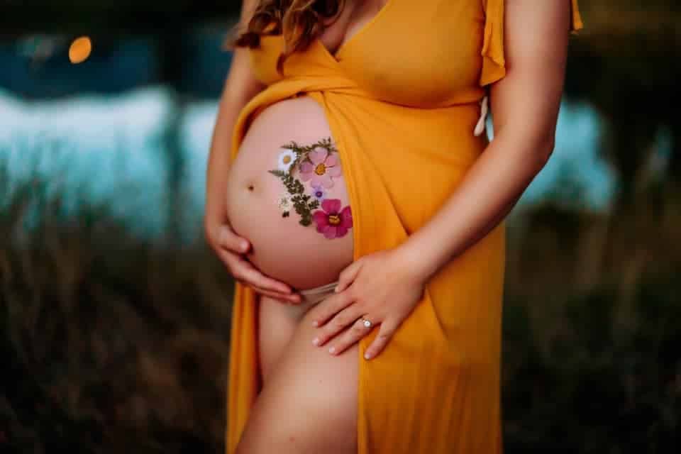 A pregnant woman in a yellow dress has flowers painted on her belly.