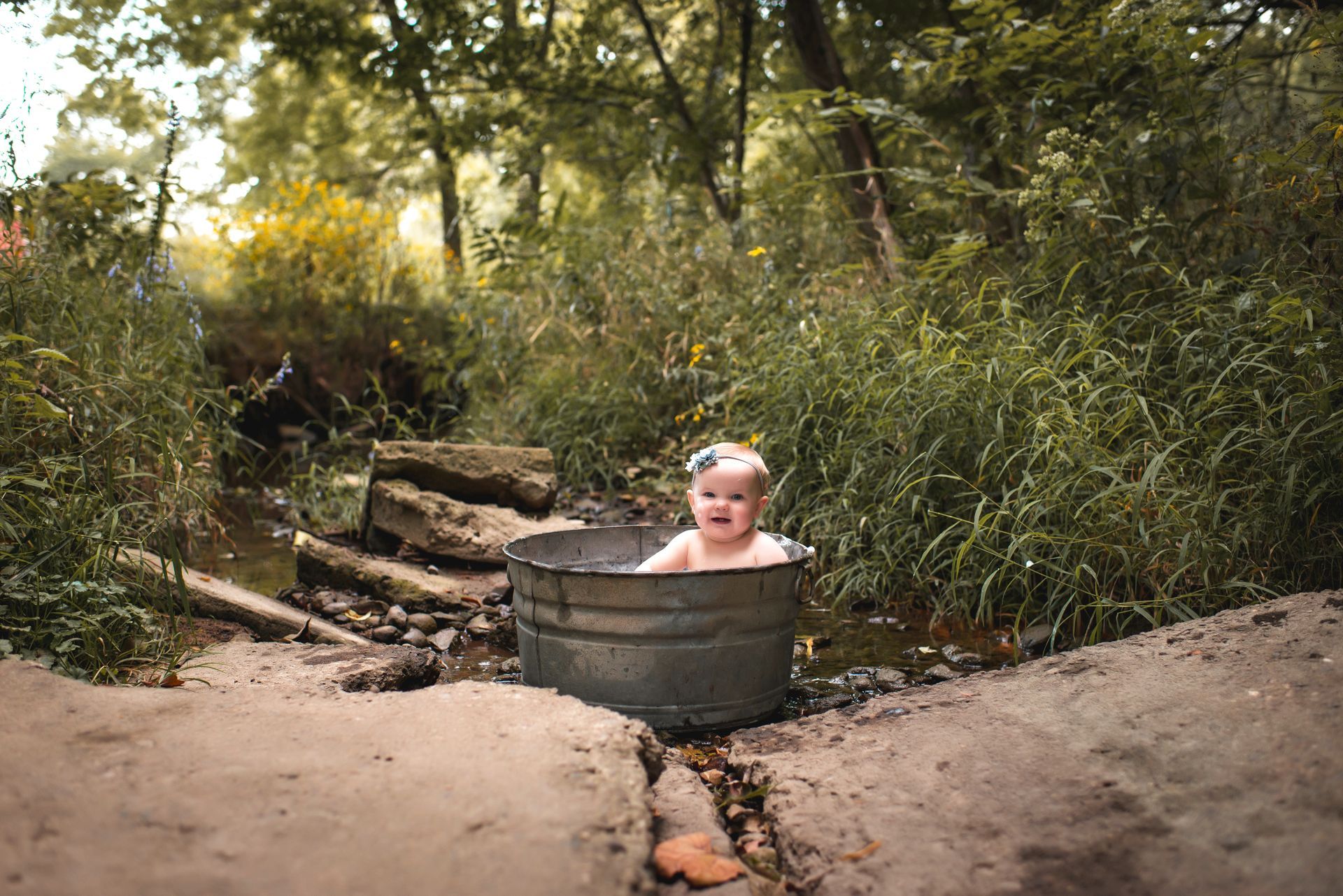 A baby is sitting in a metal tub in the woods.