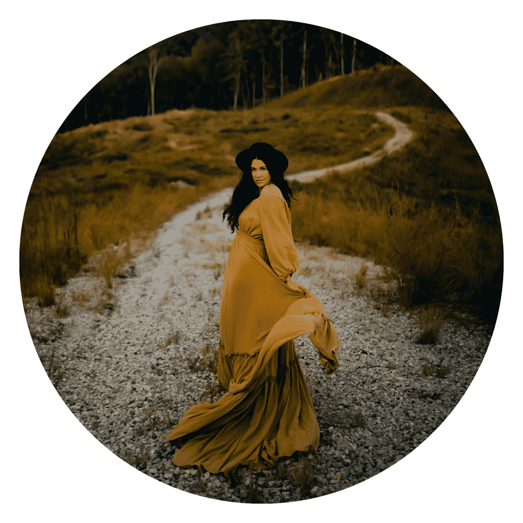 A woman in a long yellow dress is standing on a dirt road.