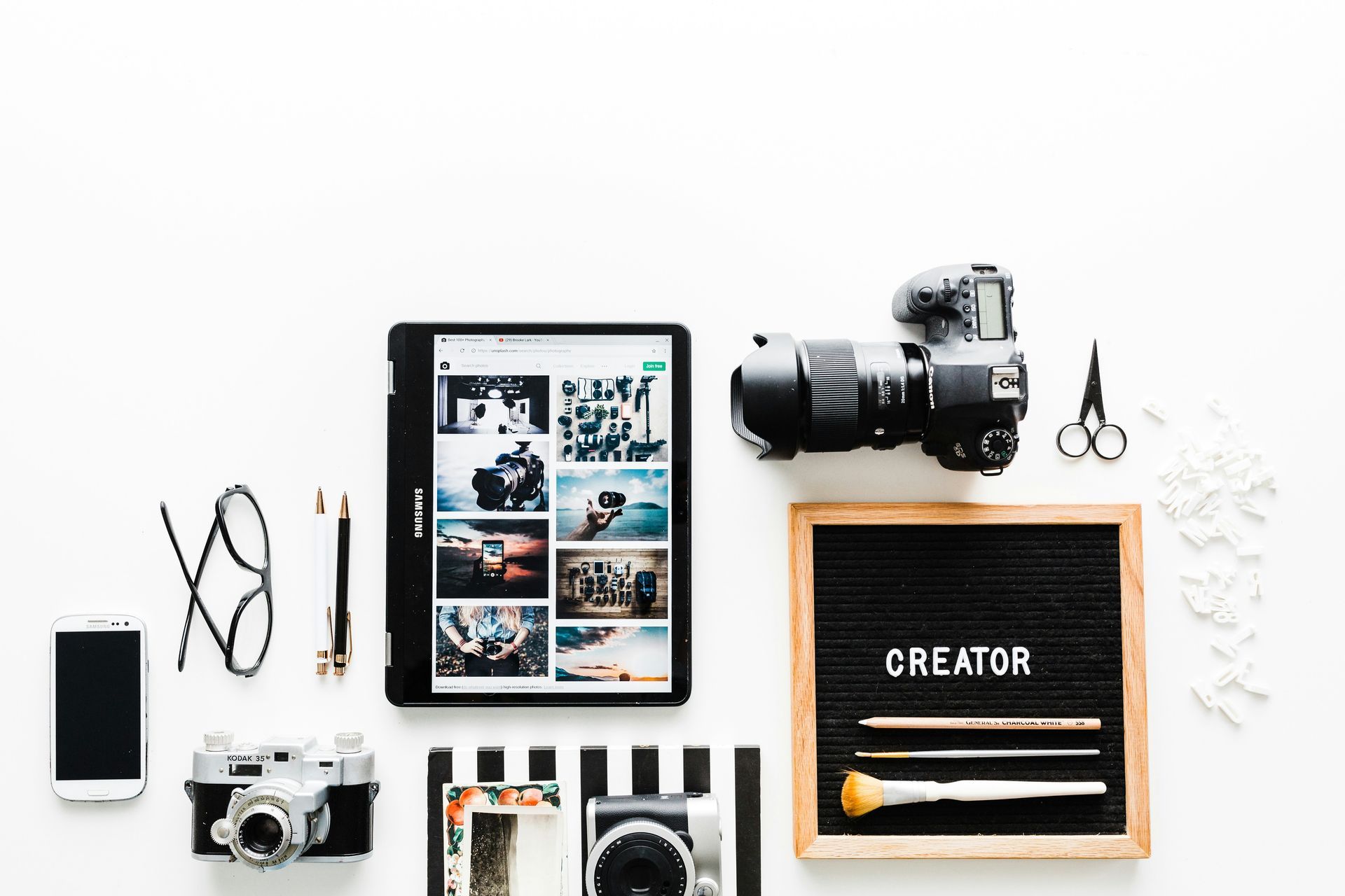 A tablet , camera , cell phone , scissors , and a letter board are on a table.