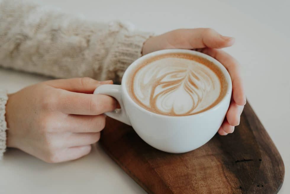 A person is holding a cup of cappuccino on a wooden cutting board.